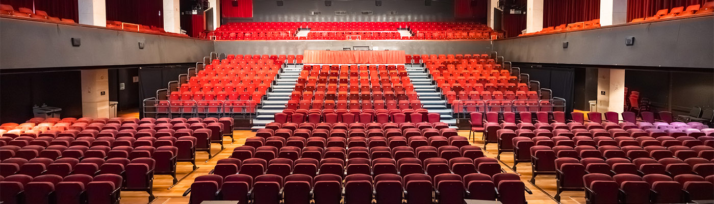 A large, empty Alban Arena auditorium with rows of red seats facing a stage with red curtains. The seating area is divided into an upper and lower section, separated by aisles and a staircase in the middle. The lighting is bright, illuminating the entire space.