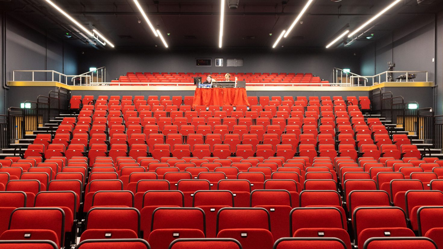 A modern Eric Morecambe Centre theatre auditorium with rows of red seats. The seats are neatly arranged, facing the stage. There are two levels, with the lower level containing the majority of the seating. The upper level contains a small balcony area with fewer seats.