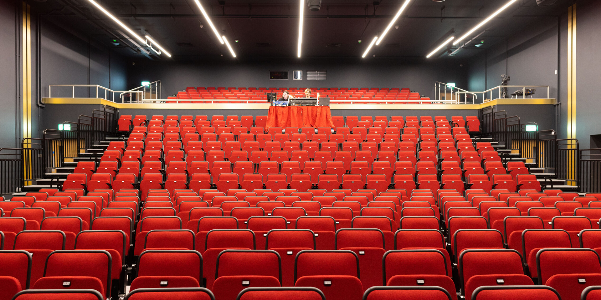 A modern Eric Morecambe Centre theatre auditorium with rows of red seats. The seats are neatly arranged, facing the stage. There are two levels, with the lower level containing the majority of the seating. The upper level contains a small balcony area with fewer seats.