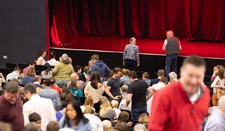 A crowded theater audience is seated in anticipation. Two people are seen walking towards the stage.