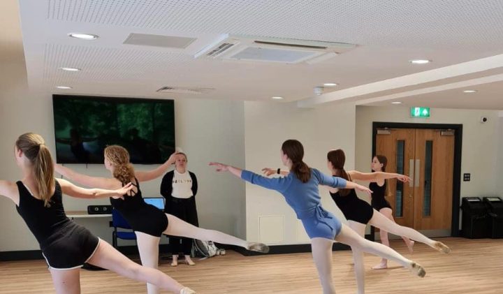 A group of ballet dancers in a studio practice an arabesque pose, with one leg extended behind them.