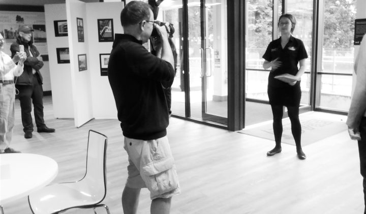A person is taking a photo of another individual speaking to a small group in a gallery setting.
