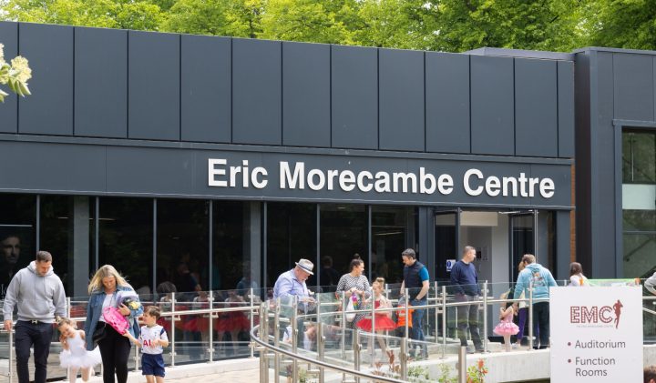 People walking in and out of a building labelled "Eric Morecambe Centre." The sign on the right reads "EMC: Auditorium, Function Rooms, Café/Bar, Box Office." The scene is lively with adults and children, some holding balloons, and greenery in the background.