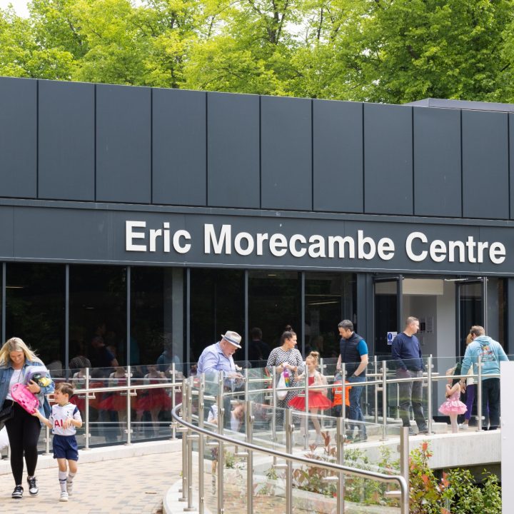 People walking in and out of a building labelled "Eric Morecambe Centre." The sign on the right reads "EMC: Auditorium, Function Rooms, Café/Bar, Box Office." The scene is lively with adults and children, some holding balloons, and greenery in the background.