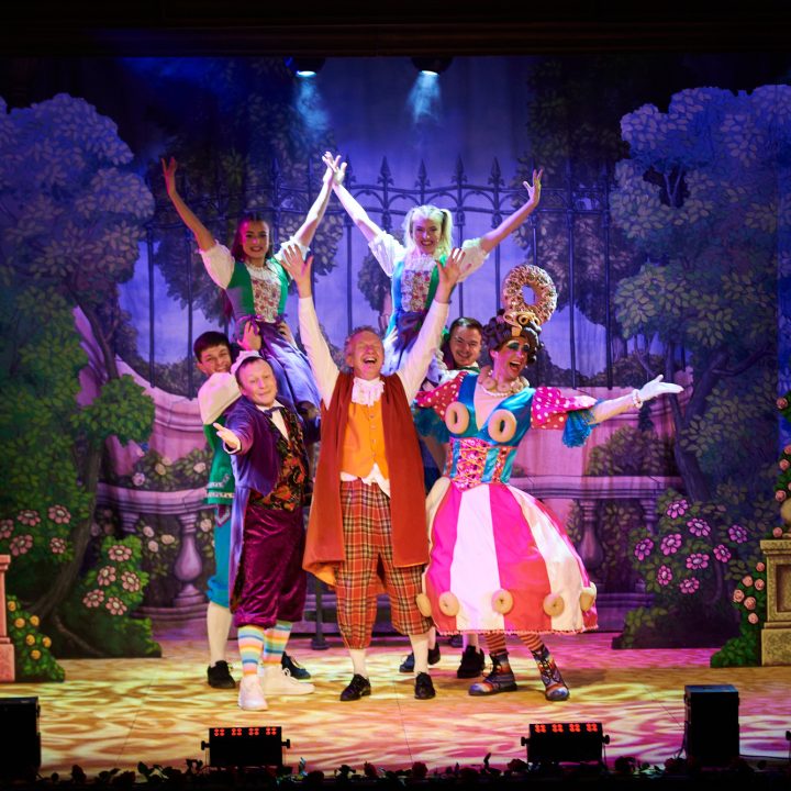 A lively stage production with vibrant costumes features six performers standing in various poses. The backdrop is a whimsical garden scene with lush greenery and flowers. The performers are dressed in colourful, eccentric outfits, and the stage is well-lit with spotlights.