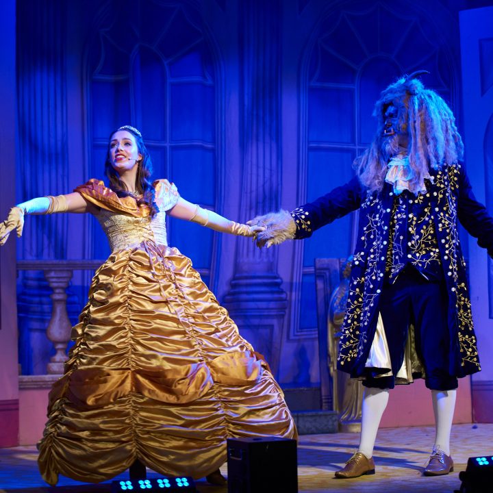 Two actors in an elaborately designed stage set, both in detailed costumes. The actor on the left is dressed as Belle in a yellow ball gown, and the actor on the right is dressed as the Beast in a blue ornate suit and mask, reminiscent of the characters from "Beauty and the Beast".