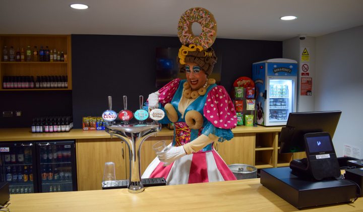 A person dressed in a colourful, whimsical costume with large pastries on their head is behind a bar, pouring a drink from a tap. The bar has various bottled drinks and snacks on shelves, a refrigerator, and a cash register. The setting appears fun and lively.