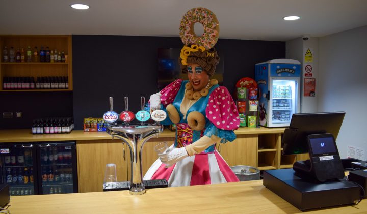 A person dressed in a colourful, whimsical costume with large pastries on their head.