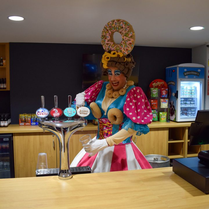 A person dressed in a colourful, whimsical costume with large pastries on their head is behind a bar, pouring a drink from a tap. The bar has various bottled drinks and snacks on shelves, a refrigerator, and a cash register. The setting appears fun and lively.