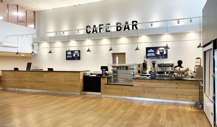 Spacious Gordon Craig café bar with light wooden floors and a high ceiling. The counter is long with wooden panelling, featuring a pastry display, coffee machines, and various beverages. The words "Café Bar" are prominently displayed on the wall above the counter.