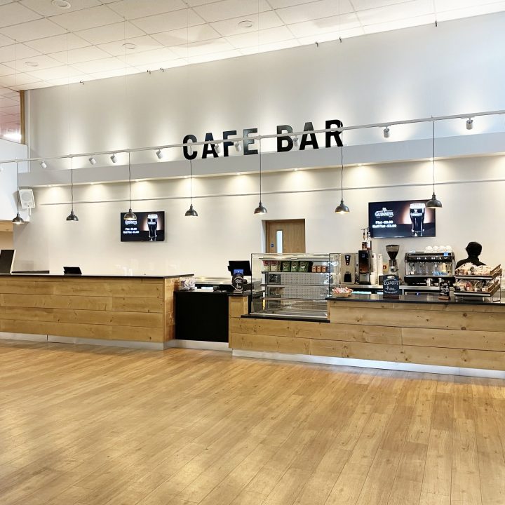 Spacious Gordon Craig café bar with light wooden floors and a high ceiling. The counter is long with wooden panelling, featuring a pastry display, coffee machines, and various beverages. The words "Café Bar" are prominently displayed on the wall above the counter.