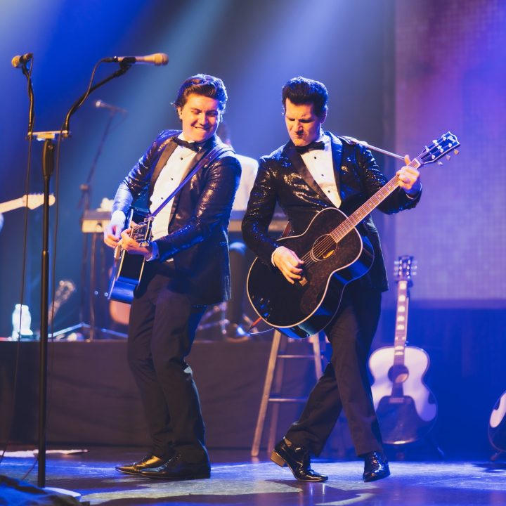 A dynamic music performance on stage features two musicians in black sequined jackets playing acoustic guitars. One looks at the other smiling. There's another guitarist in the background to the left, and several guitars rest on stands to the right. The stage is illuminated with vibrant lights.