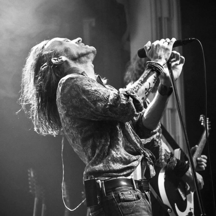 Black and white photo of a long-haired performer passionately singing into a microphone, tilting their head back with eyes closed. Wearing a patterned shirt and wristbands, they are on stage with another musician visible in the background playing a guitar.