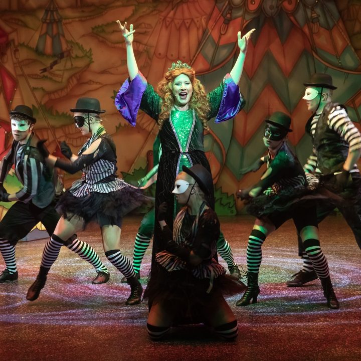 A group of performers in eccentric, colourful costumes and white face masks dance energetically on a stage with whimsical, carnival-themed backgrounds. The central figure, a woman in a vibrant green corset and black outfit, stands with her arms raised high and sings passionately.