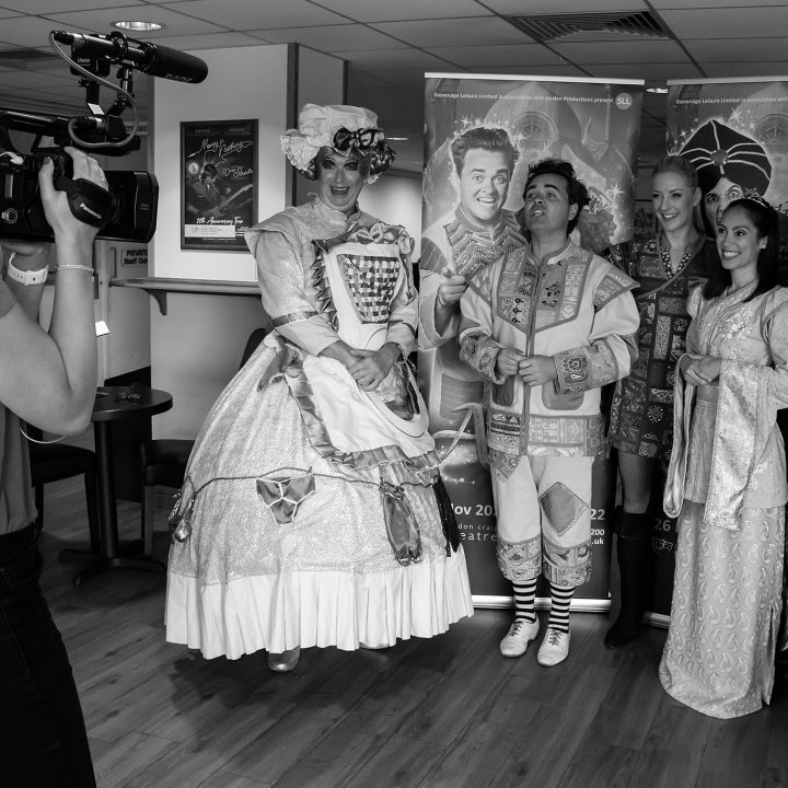 A camera operator films five people in costume. They stand in front of promotional posters and pose for the camera. The costumes include a large dress, a jester outfit, and medieval attire, suggesting a theatrical event. The setting is an indoor room with wooden floors.