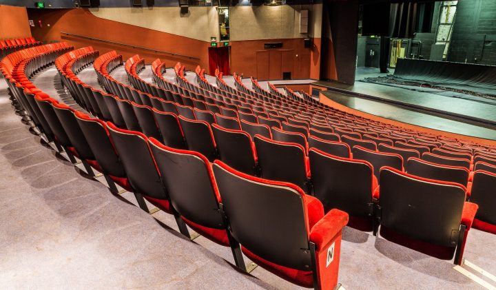 Empty Gordon Craig theatre with curved rows of black and red seats facing a stage. The stage has a closed curtain and visible stage lights hanging above. The walls and carpet are muted in colour. Scene appears well-lit with a clear view of the seating arrangement and stage setup.