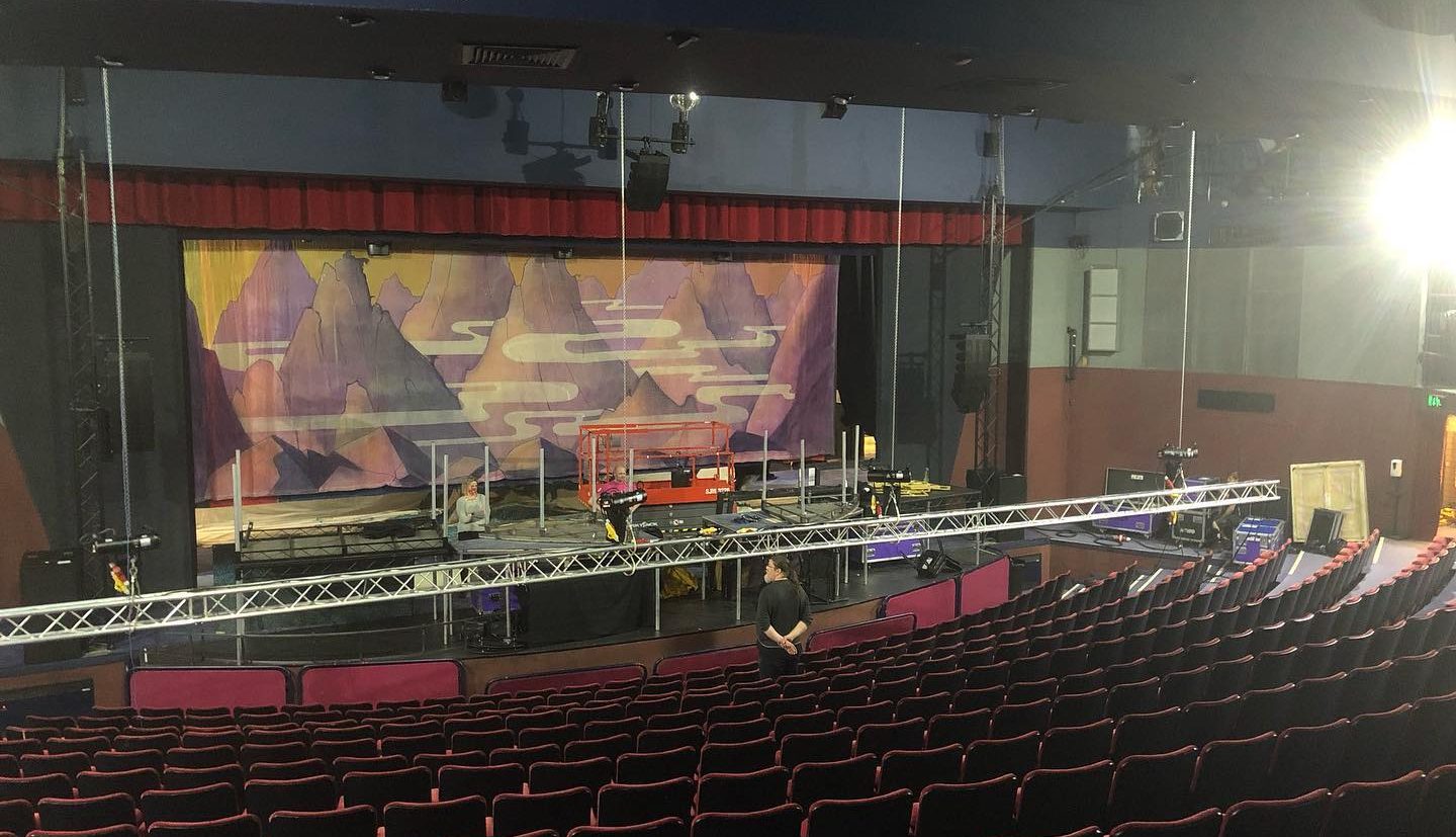 A Gordon Craig stage is under construction, with scaffolding and lighting equipment set up. The seating area is empty, and a person is standing near the front rows. The backdrop features mountainous scenery with a purple and orange colour scheme.