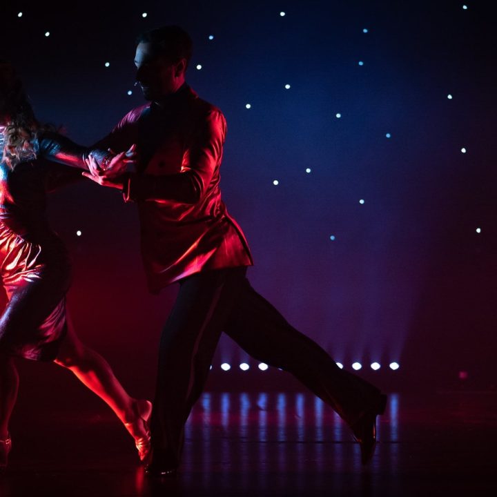 Two dancers performing on stage. The male dancer is dressed in a red shirt and black pants and is guiding the female dancer, who is wearing a shimmering red dress. The background is dark with a backdrop of scattered, glowing lights resembling stars.