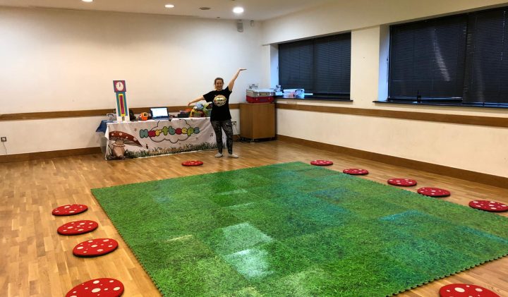 A child stands in the middle of a room with a large green mat that resembles grass, surrounded by small red cushions with white polka dots. Behind the child is a table with various colourful items and decorations on it. The room features wooden floors and large windows.