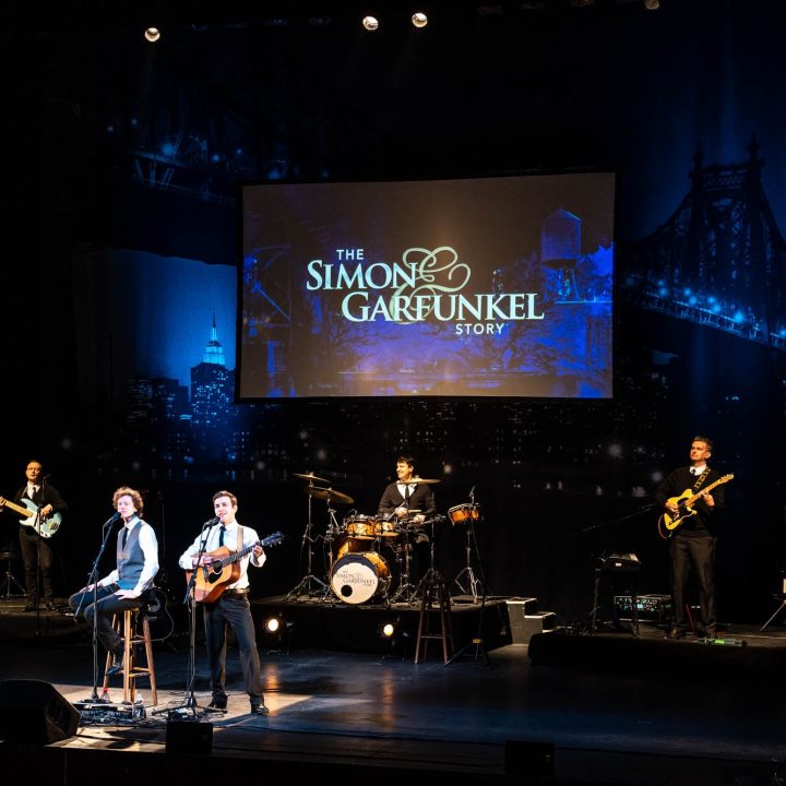 A band performs on stage with a backdrop reading "The Simon & Garfunkel Story." Two musicians sit on stools with guitars and microphones. Behind them is a drummer, keyboardist, and bassist. The stage is illuminated with spotlights, and the audience is not visible.