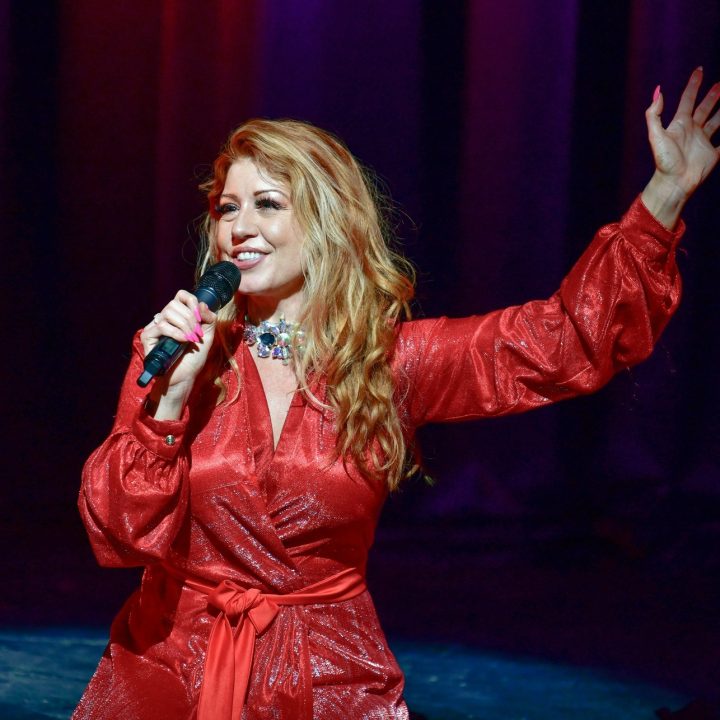 A woman with long, wavy blonde hair, wearing a shiny red dress, is holding a microphone with one hand and raising the other hand in the air. She appears to be singing on stage with a backdrop of purple lights.