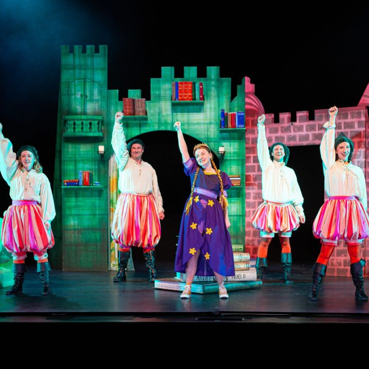 A group of five costumed performers, four in pink and white traditional attire and one in a purple dress with stars, stand on Grove Theatre stage with a castle backdrop. They are all holding one arm up in the air, appearing to be in the middle of a lively performance.