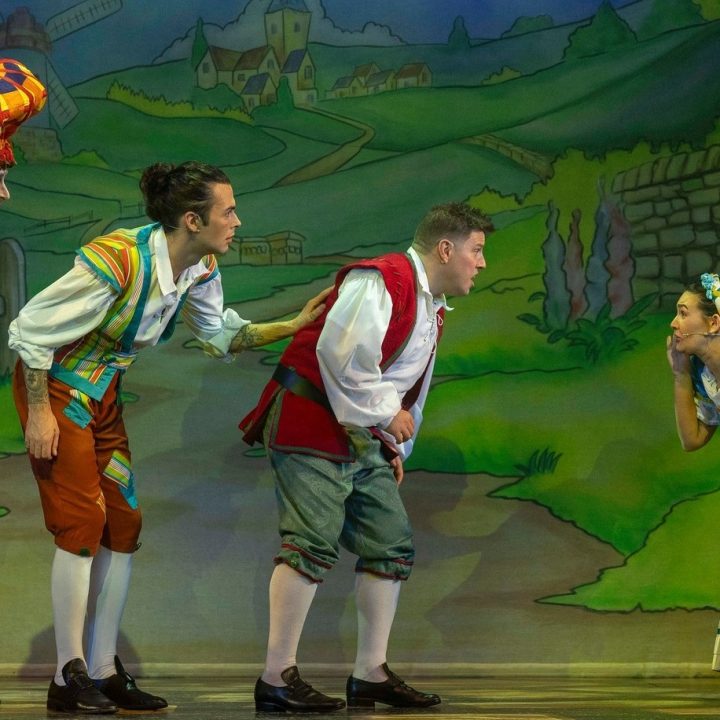 Four actors perform on Grove Theatre stage in a colourful theatrical scene. One actor in a vibrant, plaid dress and wig leans towards another in historical attire, who appears to whisper to the next actor. A fourth actor in a blue dress blows a kiss. The backdrop depicts a village.