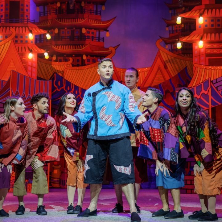 A group of performers in colourful, patchwork costumes stands together on Grove Theatre stage, with an ornate backdrop featuring vibrant pagoda-style buildings and warm lighting. The central figure, in a blue shirt, appears to be leading or singing, as others look towards him.