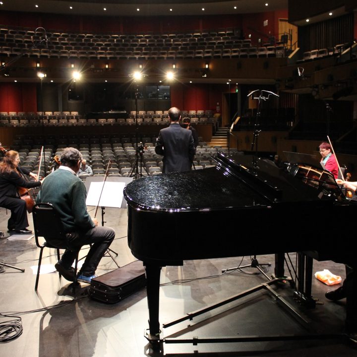 A group of musicians, including a pianist, violinist, and cellist, are rehearsing on stage in an empty Grove Theatre. The stage is equipped with music stands, microphones, and a grand piano. The conductor stands at the front, facing the musicians.