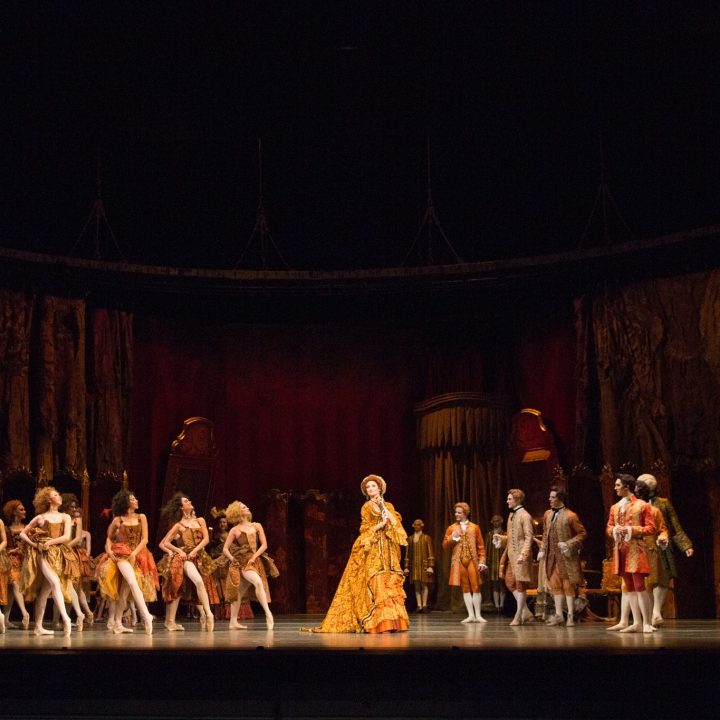 A stage performance featuring a group of ballet dancers in elegant costumes at The Grove Theatre. The central figure is a person dressed in an elaborate, golden outfit. The background consists of dark, ornate set pieces with classical and regal elements, creating a majestic atmosphere.