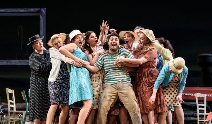 A group of people on Grove Theatre stage, many wearing hats and vintage clothing, surround a man in a striped shirt and suspenders, who appears to be singing or shouting gleefully. The scene is lively, with chairs and set pieces visible in the background.