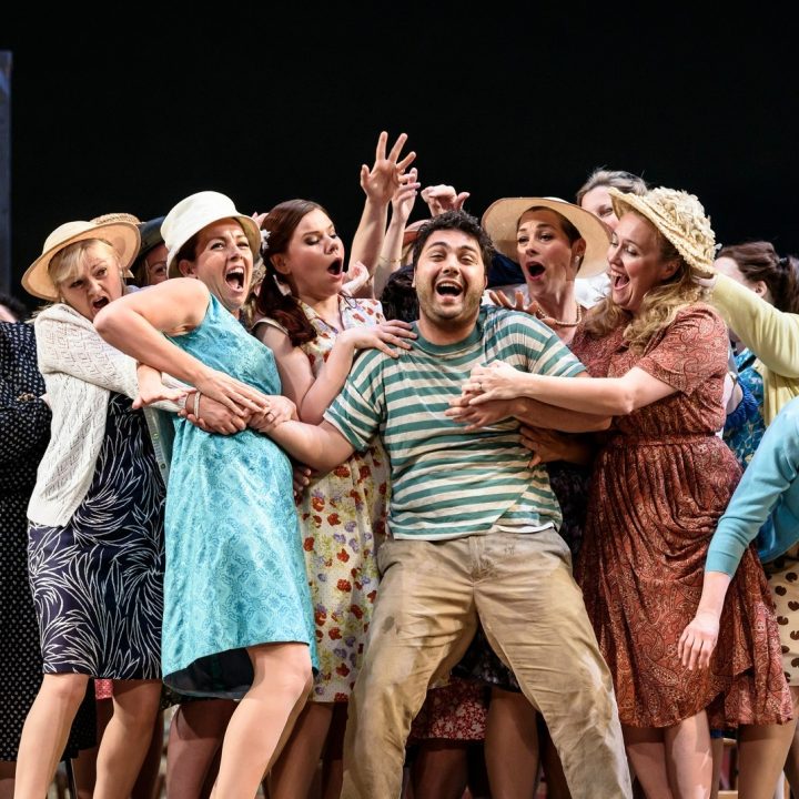 A group of people on Grove Theatre stage, many wearing hats and vintage clothing, surround a man in a striped shirt and suspenders, who appears to be singing or shouting gleefully. The scene is lively, with chairs and set pieces visible in the background.