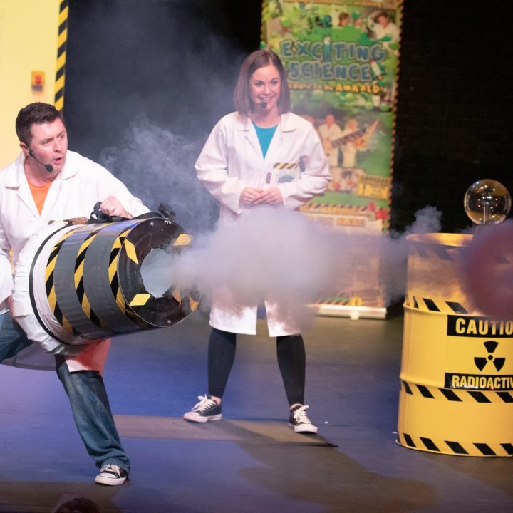 Two people in white lab coats perform a science experiment. The person on the left is holding a smokey barrel with caution tape, and the person on the right is smiling while observing. Various science props and safety symbols are in the background.