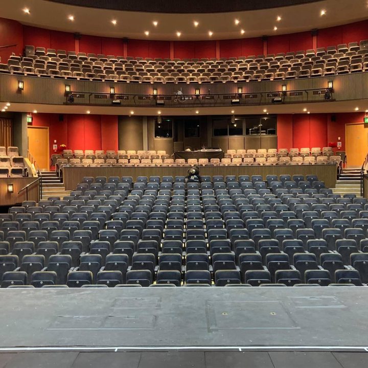 A large, empty Grove Theatre with rows of black seats and balconies on either side, all facing a stage. The theatre is well-lit, with a person sitting alone in the centre row of seats, surrounded by unoccupied seats. Bright lights line the ceiling and walls.