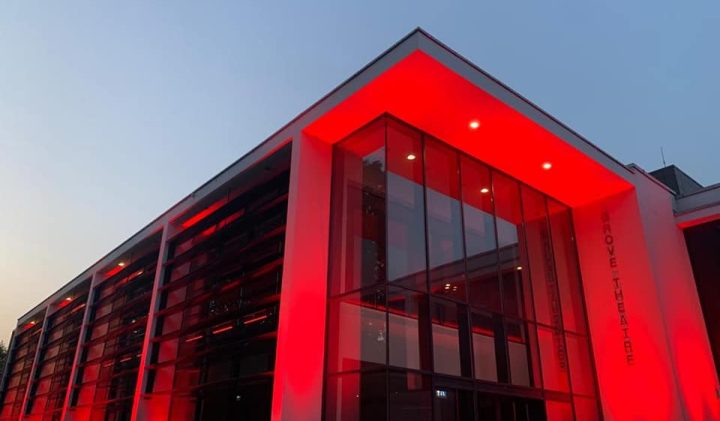 A modern Grove Theatre Gallery building, the exterior of which is illuminated with red lights. The structure features large glass windows and a sleek, clean design with prominent vertical and horizontal lines. The sky is clear with a hint of dusk.