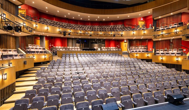 A wide-angle view of a modern, empty Grove Theatre auditorium. The seating consists of plush, light grey chairs arranged in rows on the main floor and two balconies. Warm lighting and wooden accents adorn the space, giving it a cosy yet elegant atmosphere.