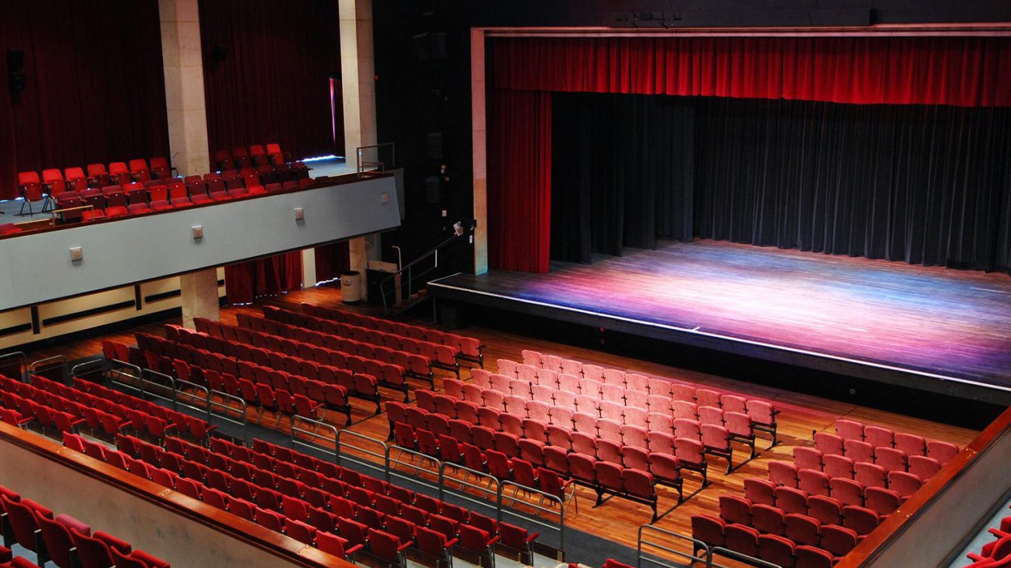 A spacious, empty Alban Arena theatre with rows of red seats facing a large stage. The stage is framed by red curtains and illuminated from above. There are balconies with additional seating on both sides of the main seating area. The theatre has a modern design.