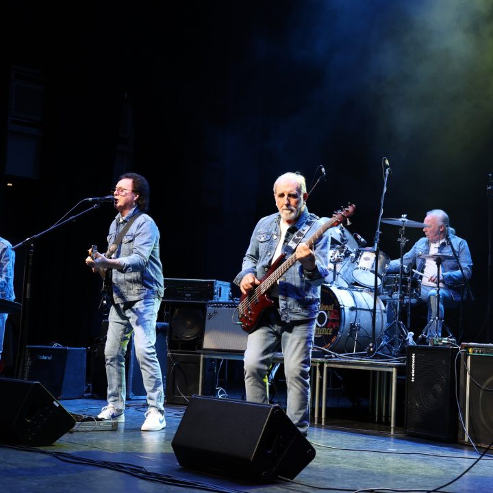 A rock band performs on stage, with four older male musicians passionately playing their instruments. One is on keyboard, another on guitar, a third on bass guitar, and a fourth on a drum set. They are dressed in denim jackets and jeans, with colourful stage lighting.