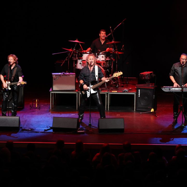 A rock band performs on stage with bright lighting. Four members are visible: one playing keyboard, one playing bass guitar and singing into a microphone, one playing electric guitar, and one playing drums in the background. Amplifiers and equipment are also on stage.