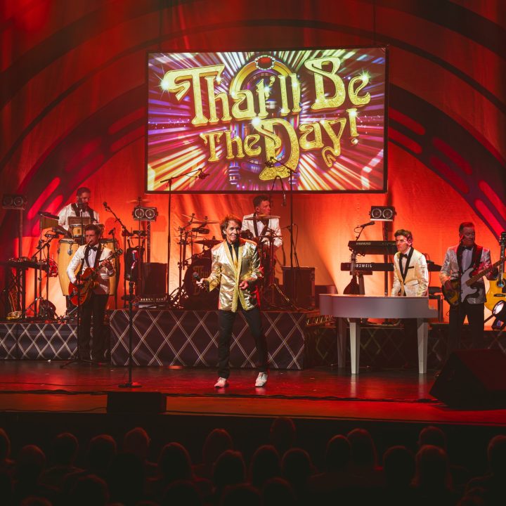 A live band performs on stage with vibrant red lighting and a colourful backdrop displaying the words "That'll Be The Day!" Band members play various instruments including guitars, keyboards, and drums. The audience appears as silhouettes at the bottom of the image.