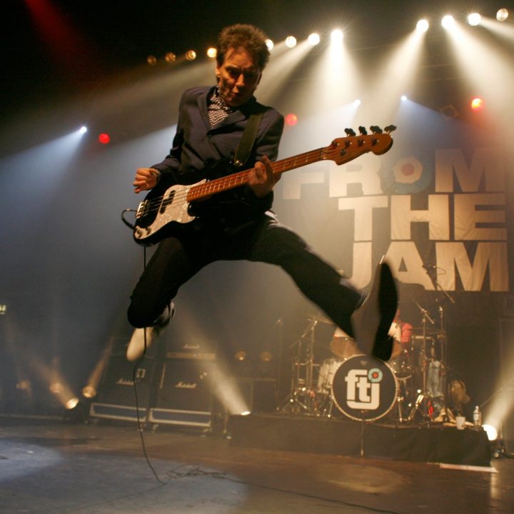 A musician dressed in a dark suit jumps in mid-air while playing a guitar on stage, illuminated by stage lights. Another musician plays guitar to the left. The backdrop displays the text "FROM THE JAM" with a stylized "ftj" logo on the drum kit.