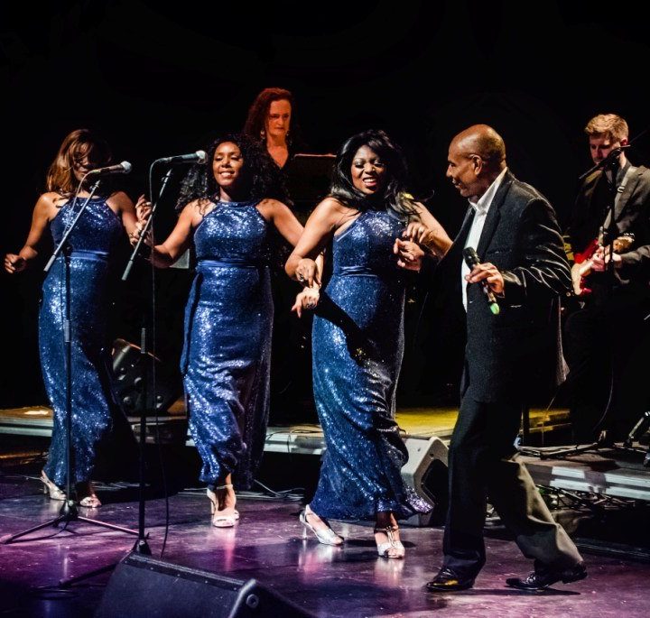 A band is performing on Alban Arena stage. Three women in matching blue sequin dresses are singing into microphones while dancing. A man in a suit is dancing beside them. In the background, musicians are playing instruments, including a drummer and a guitarist.