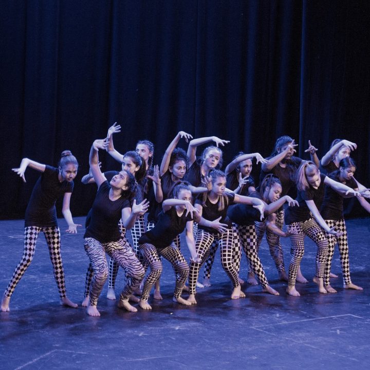 A group of young dancers, wearing black tops and patterned leggings, perform a contemporary dance routine on a dimly lit stage. They are positioned in a cohesive formation, with their arms extended and expressive poses conveying movement and emotion.