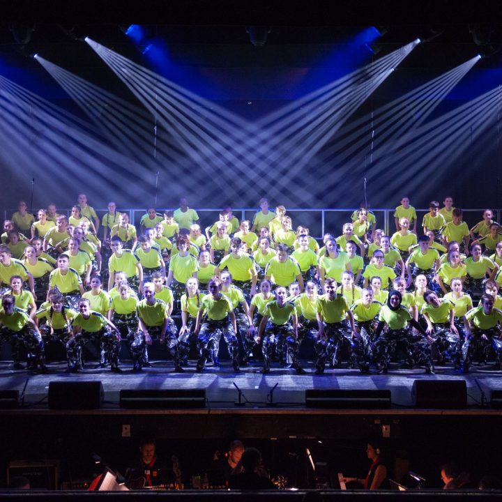 A large choir wearing matching bright green shirts performs on stage under a series of spotlights. The choir members are arranged in several rows on risers, and musicians in the orchestra pit are partially visible at the bottom of the image. The background is dark.