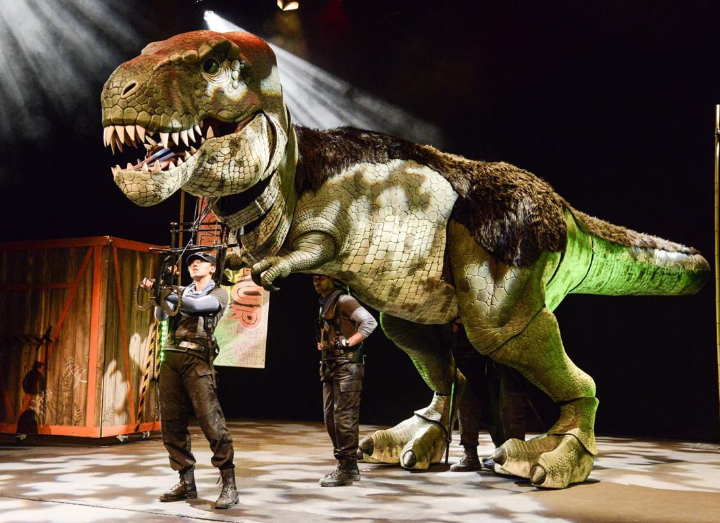 A large, realistic T-rex puppet is featured on Alban Arena stage with two visible puppeteers and a performer in front. The scene is brightly lit with spotlights, and the background includes stage props and banners. The setting appears to be a theatrical dinosaur show.