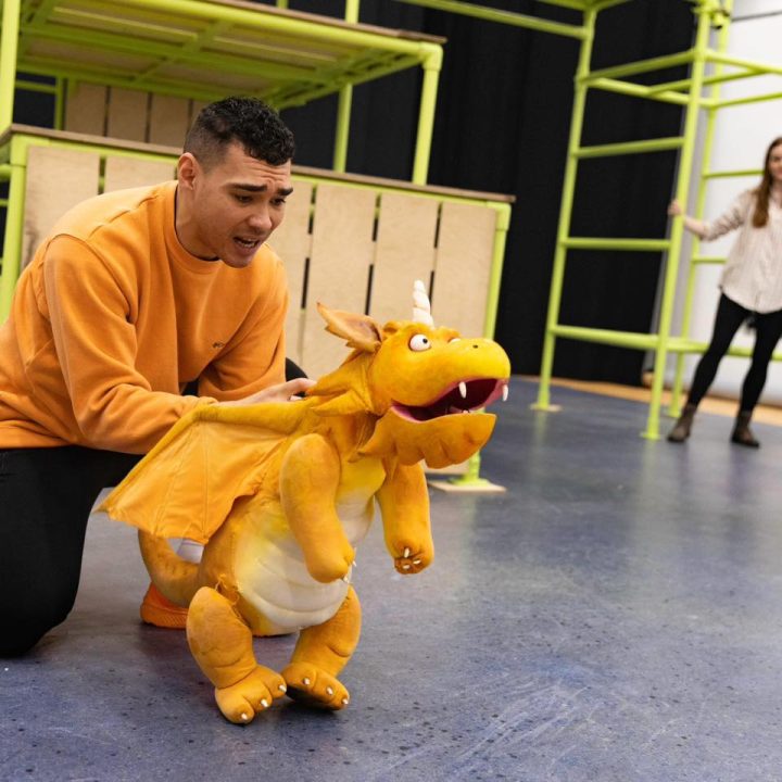 A person kneels on the floor while holding a small, orange dragon puppet with an open mouth. In the background, a second person stands near green climbing frames. The setting appears to be an indoor play area or rehearsal space.