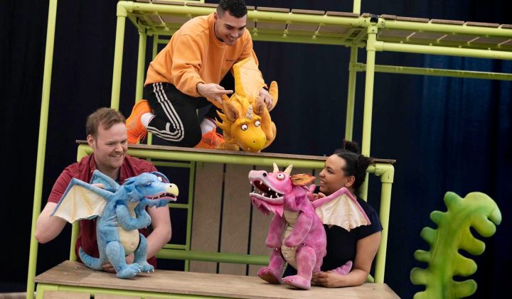 Four people are interacting with colourful dragon puppets on an elaborate stage setup. Two individuals are on an upper level with an orange dragon, while the other two are at a lower level with blue and pink dragons. Green stage props are in the foreground.