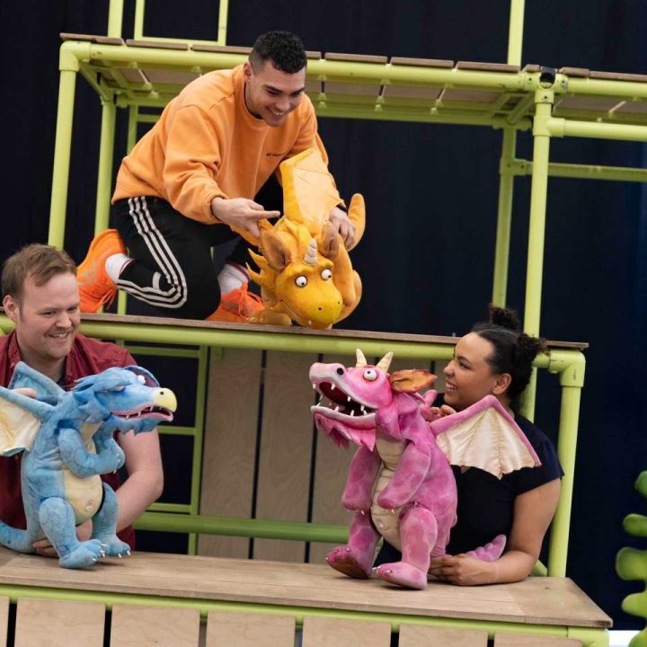 Four people are interacting with colourful dragon puppets on an elaborate stage setup. Two individuals are on an upper level with an orange dragon, while the other two are at a lower level with blue and pink dragons. Green stage props are in the foreground.