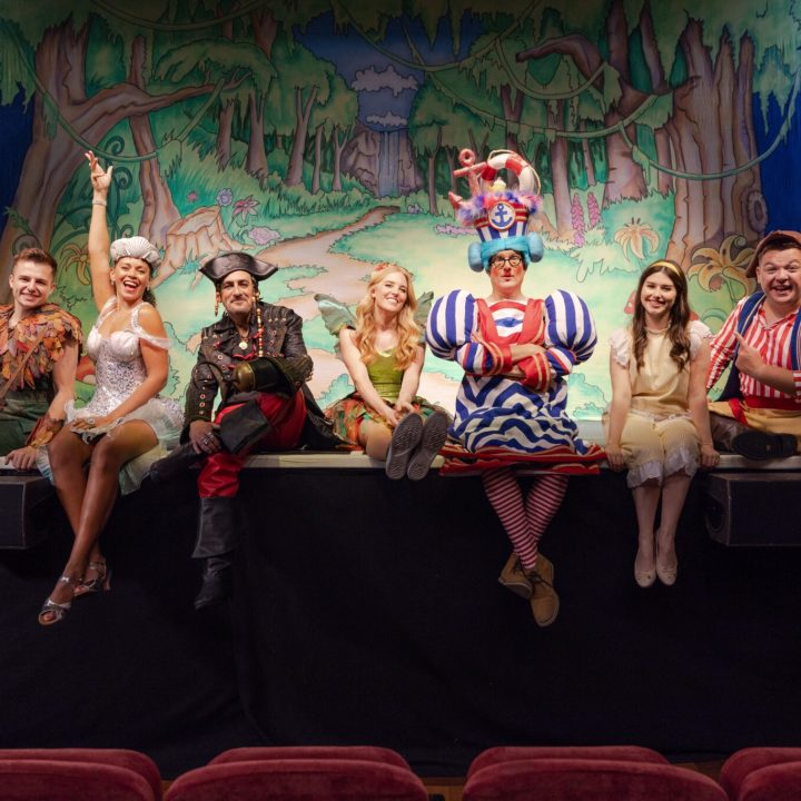 A group of people in colourful, whimsical costumes sit and stand on the edge of a stage. The background is a vibrant, painted forest scene. They are all smiling and some are striking cheerful poses, giving a lively and joyful atmosphere to the scene.