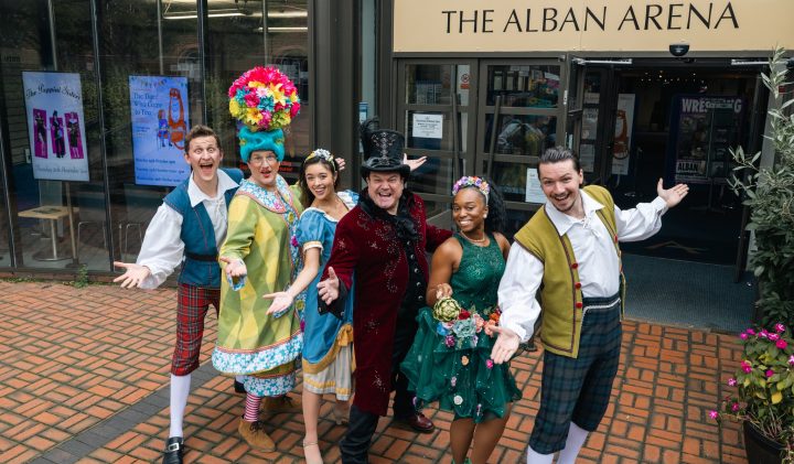 A group of six people in colourful, theatrical costumes stand happily with arms open in front of The Alban Arena. They are smiling and standing on a brick pavement near a flowerbed, seemingly promoting a performance or event at the venue.